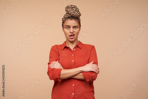 Studio photo of bewildered young brunette lady with headband grimacing confusedly her face while looking at camera and cossing hands on chest, isolated over beige background photo