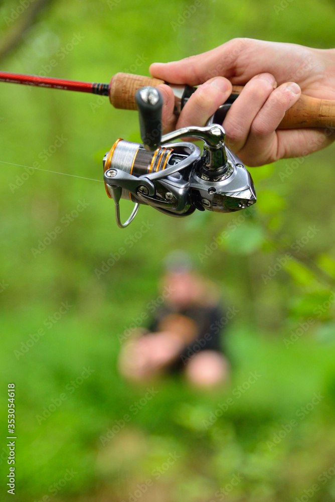 Fishing reel on rod in fisherman's hand on green background with defocused fisherman in background Fishing reel on rod in fisherman's hand on green background with defocused fisherman in background