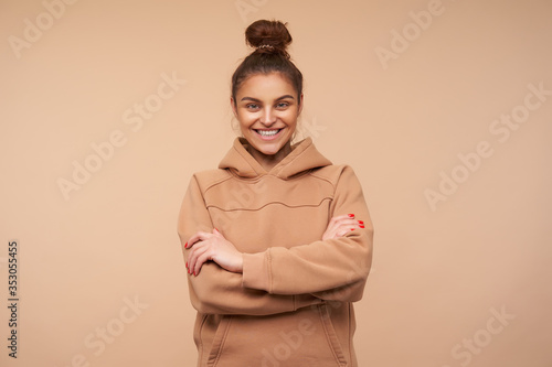 Good looking young happy brown haired lady showing her white perfect teeth while smiling cheerfully and crossing hands on her chest, posing over beige background © timtimphoto