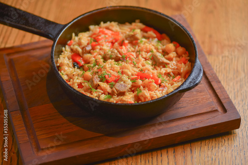 Rice with meat and vegetables served in a frying pan on wooden board over light rustic wooden table