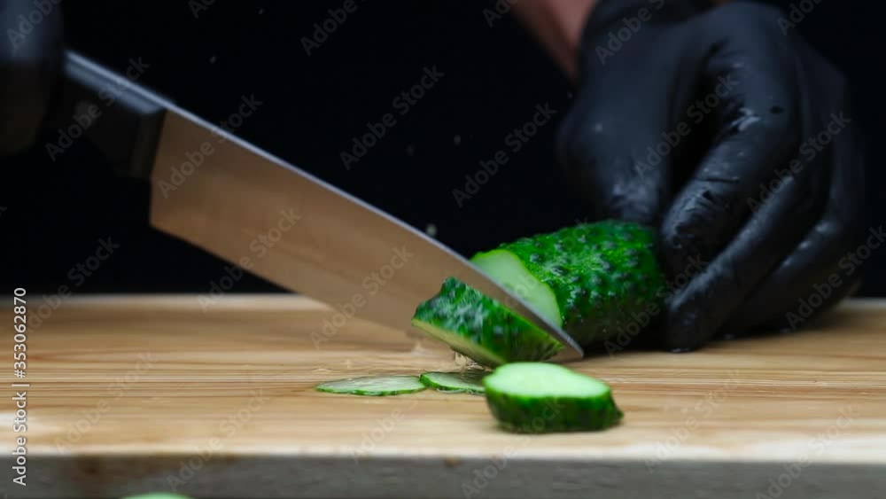 The cook in the kitchen in black gloves cuts cucumbers with a knife. The footage was shot in slow motion