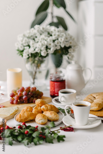 Hearty breakfast of mini croissants, pastry, coffee in cups, grapes and jam in a jar, served on white table, decorated with white flowers bouquet, candles and sprig with green leaves and red berries
