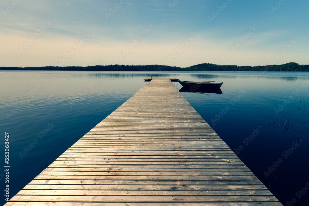 A wooden boat with oars moored to a pier on a lake in spring. The pier from the planks leaves with the prospect towards the horizon. The sun in the blue sky is approaching sunset. Latvia