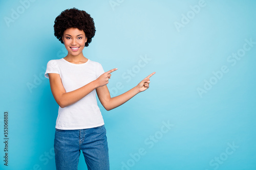 Photo of beautiful dark skin curly lady toothy smiling indicating fingers empty space proposing shopping low sale prices wear casual white t-shirt jeans isolated blue color background