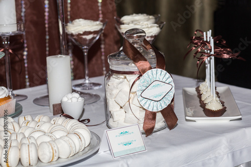 Vanilla macaroons on a round dish, creamy marmalade in a glass jar, tiramisu in glasses and cake on sticks, shot from the side under pulsed lighting. photo