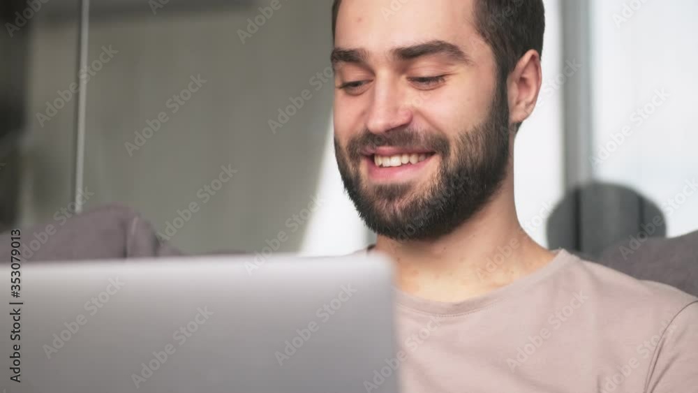 A close-up view of a happy young man is using his laptop sitting at home on the sofa in the living room A close-up view of a happy young man is using his laptop sitting at home on the sofa in the living room