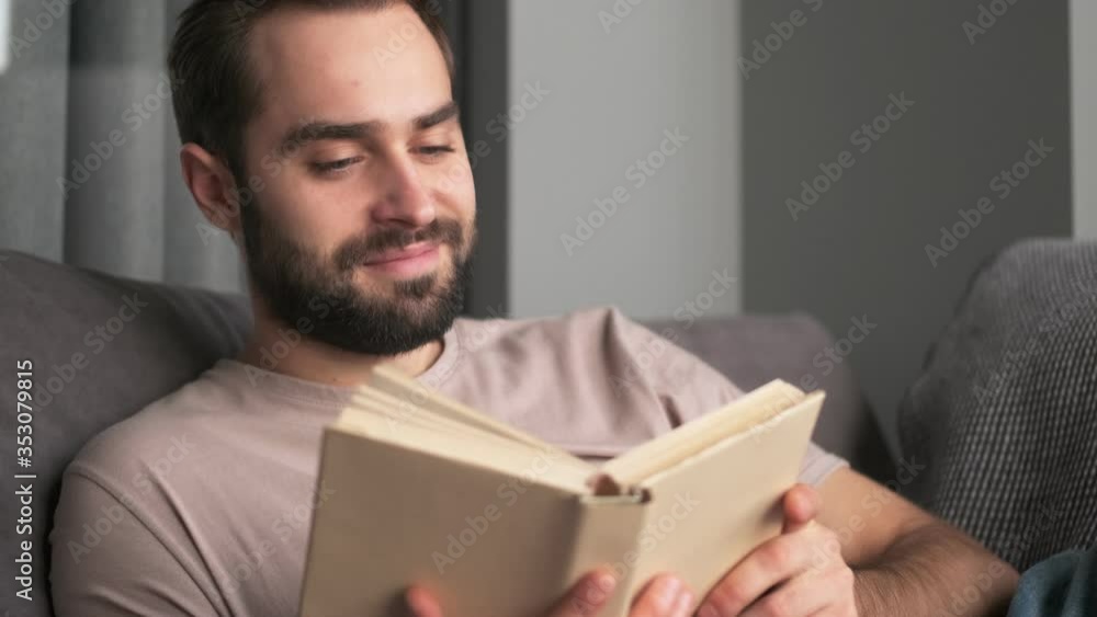 An attractive positive young man is reading a book sitting at home on the sofa in the living room An attractive positive young man is reading a book sitting at home on the sofa in the living room