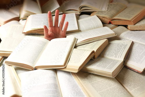 A student or reader drowned in a mountain of books. Man's hand sticks out from the rubble of open books. Concept - preparing for exams or excess information. Selective focus.