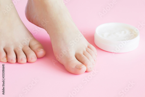 Young, perfect groomed woman feet. Opened white jar of natural herbal cream on pastel pink background. Care about clean, soft and smooth skin. Healthcare concept. Closeup.