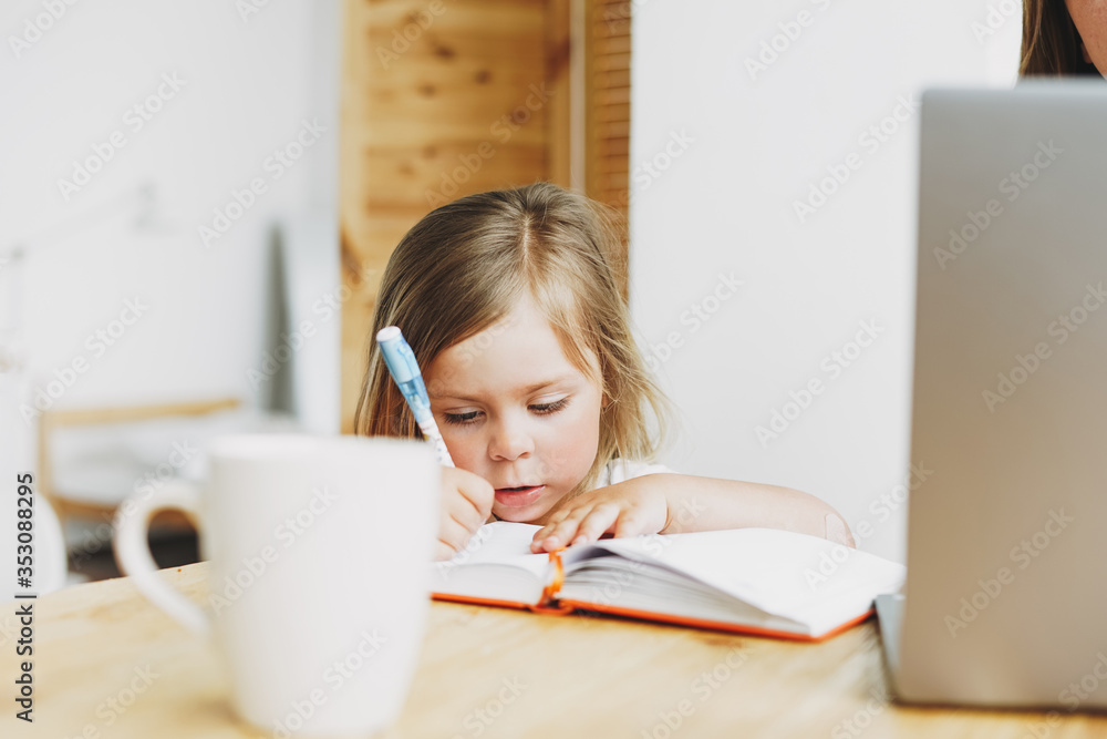 Cute toddler girl writing drawing on table with working parent near laptop at the home Cute toddler girl writing drawing on table with working parent near laptop at the home