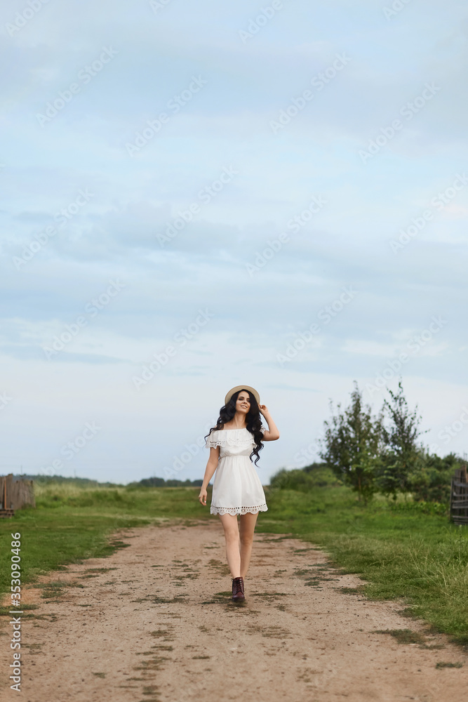 Beautiful girl in white summer dress touching straw hat and smiling while standing on rural road and looking away. Young brunette woman in short white dress walking by a rural road. Summertime