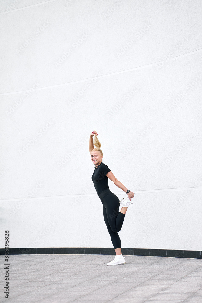 Young cute funny slim woman wearing black sportswear and trendy white sneakers have fun and posing at camera. Outdoor shot on urban white wall background. Young cute funny slim woman wearing black sportswear and trendy white sneakers have fun and posing at camera. Outdoor shot on urban white wall background.