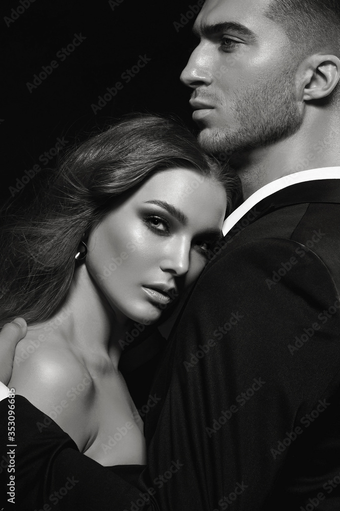 Beautiful young stylish couple, man and woman, in black evening dresses in each other's arms posing in the studio on a black background. Beautiful young stylish couple, man and woman, in black evening dresses in each other's arms posing in the studio on a black background.