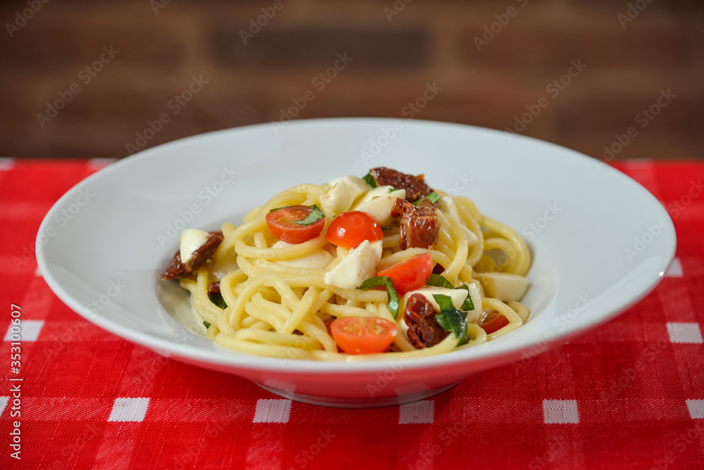 Spaghetti with cheese, dried and fresh tomatoes on white plate on table with red tablecloth Spaghetti with cheese, dried and fresh tomatoes on white plate on table with red tablecloth