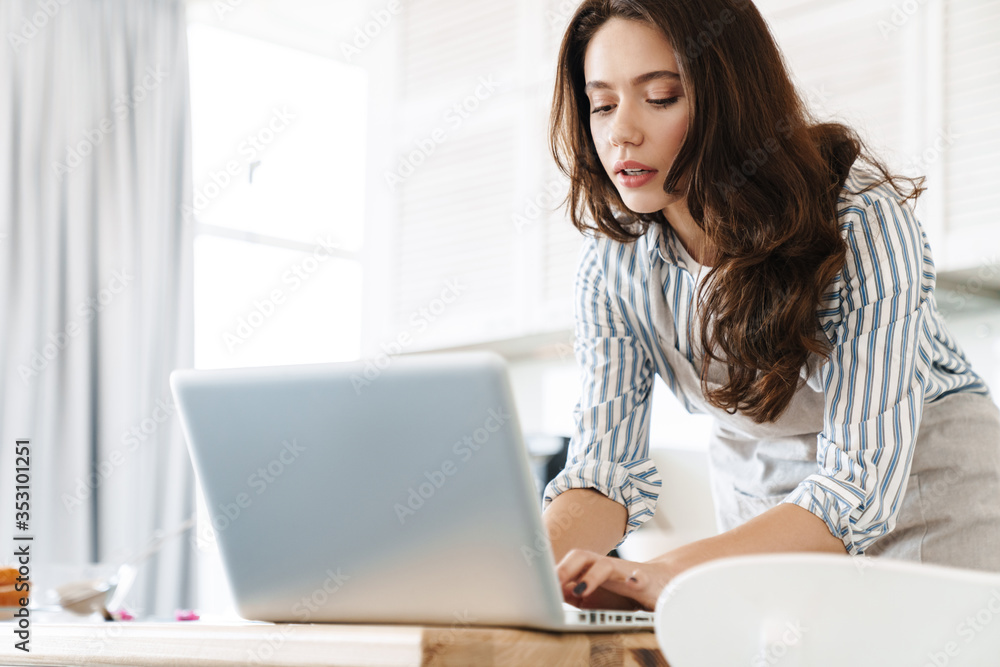 Image of thinking brunette woman using laptop while cooking pie Image of thinking brunette woman using laptop while cooking pie