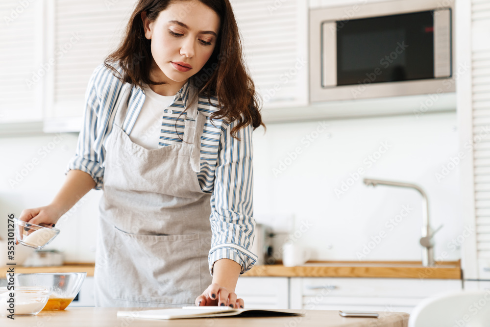 Image of focused young woman reading recipe while cooking pie