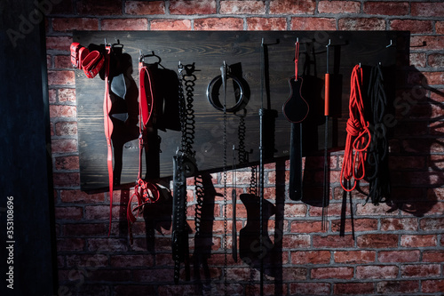 A set of varied leather lashes for sexual pleasures hang on a wooden hanger on a brick wall. Sex equipment in red and black for role-playing games. BDSM room.