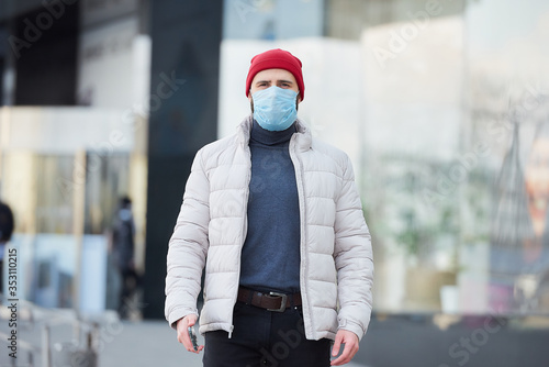 A man in a red watch cap wearing a medical face mask to avoid the spread coronavirus (COVID-19). A guy with a surgical mask on the face because of the pandemic walking in the center of the city.