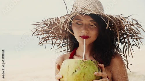 Beautiful pretty asian woman in hat and red dress drinking coconut on tropical paradise beach. Woman quench her thirst with coco nut. Beauty, baunty, travel, fashion concept. Vacation on exotic island photo