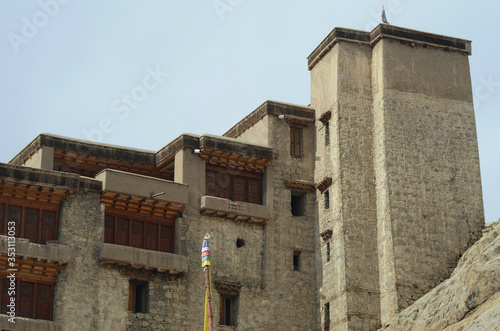 Details of the grey stone and wooden doors and windows of Leh Palace in Ladakh, India. There is a tower at the far end. A prayer flag is in the foreground, and the sky is blue.