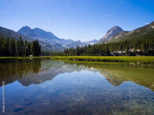 Beautiful mountain range and pine forest, reflected in a still, clear river, surrounded by alpine meadow.