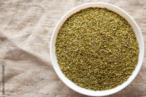 Dried Green Greek Oregano Spice in a white bowl on cloth, overhead view. Flat lay, top view, from above. Copy space.