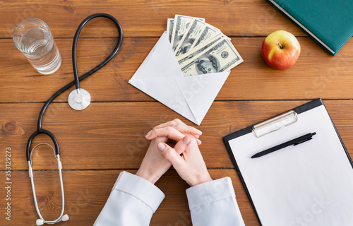 Crossed female doctor hands on wooden table with envelope with money photo