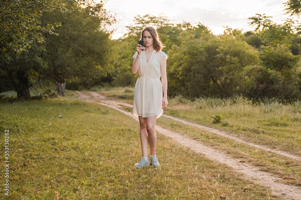 Beautiful young woman with brown hair in white dress walks through the autumn or summer forest with smartphone in her hand. Girl is looking for mobile communications and Internet in the forest. Beautiful young woman with brown hair in white dress walks through the autumn or summer forest with smartphone in her hand. Girl is looking for mobile communications and Internet in the forest.