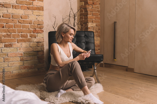 Bilde på lerret beautiful young woman sitting of shaggy carpet and chatting on smartphone at hom