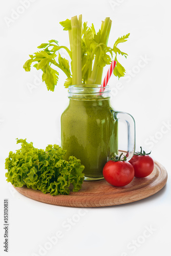 Glass mason jar of kiwi smoothie with a drinking straw standing on a round wooden kitchen shelf surrounded by leaves, a green cucumber and tomatoes about a stalk and celery leaves in the background