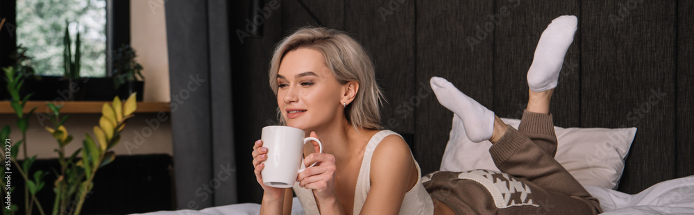 panoramic shot of attractive smiling woman holding cup of tea while relaxing on bed panoramic shot of attractive smiling woman holding cup of tea while relaxing on bed