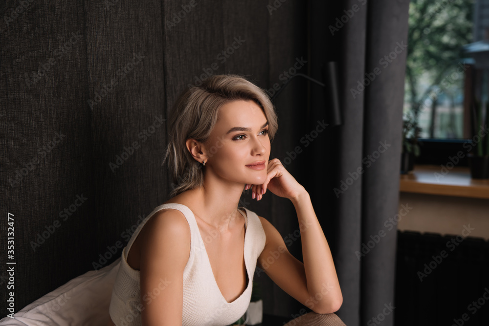 Fototapeta premium dreamy, positive woman touching face and looking away while sitting in bedroom