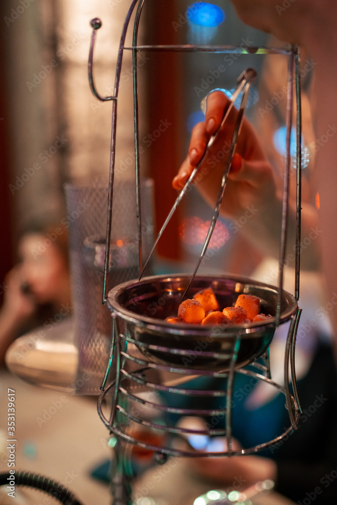 Hookah bowl with coal for smoking closeup. Hands holding forceps for shisha hookah and adjusts hot coals in metal bowl