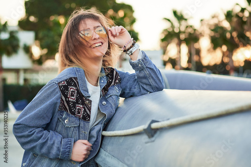 golden backlight, beautiful smile girl with long blonde hair in sunglasses, dressed in a denim jacket at summer sunset against the background of yachts in the port photo