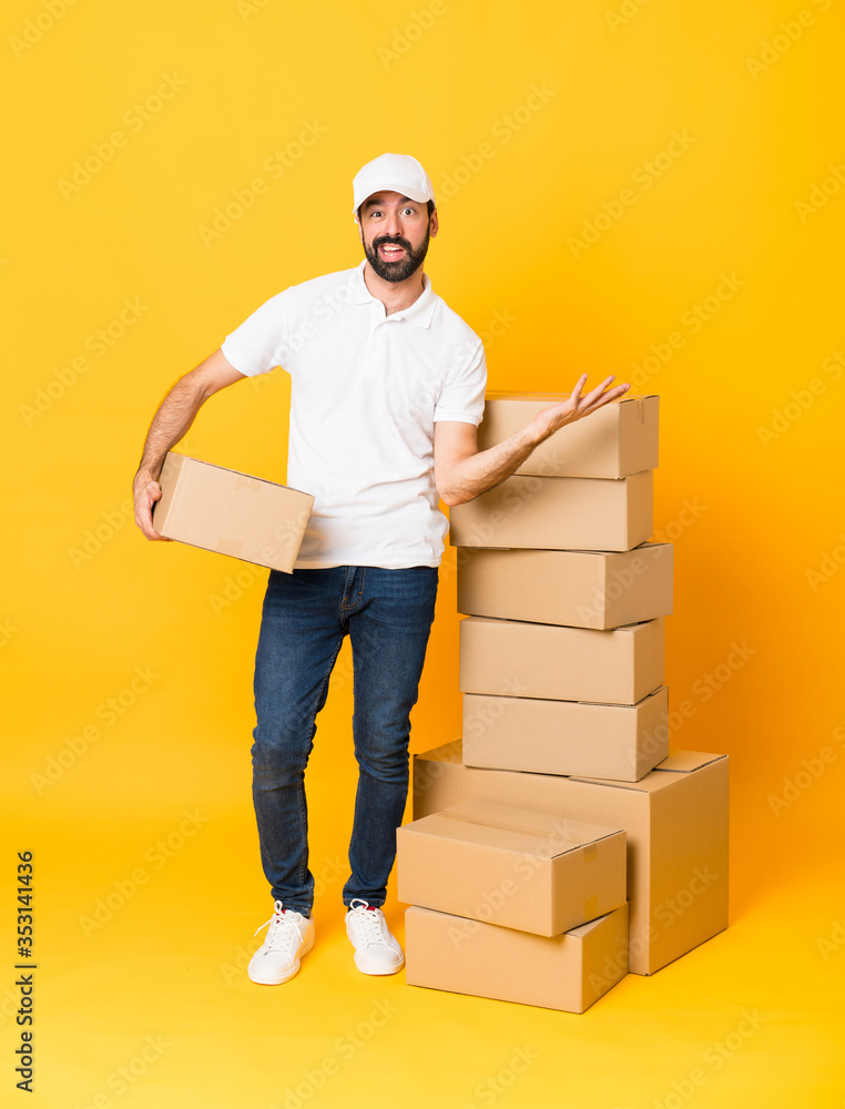 Full-length shot of delivery man among boxes over isolated yellow background making doubts gesture Full-length shot of delivery man among boxes over isolated yellow background making doubts gesture