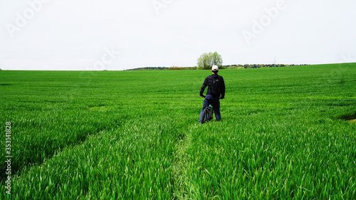 A man in a black sports jacket and a white cap on a bicycle among the green grass looks forward
