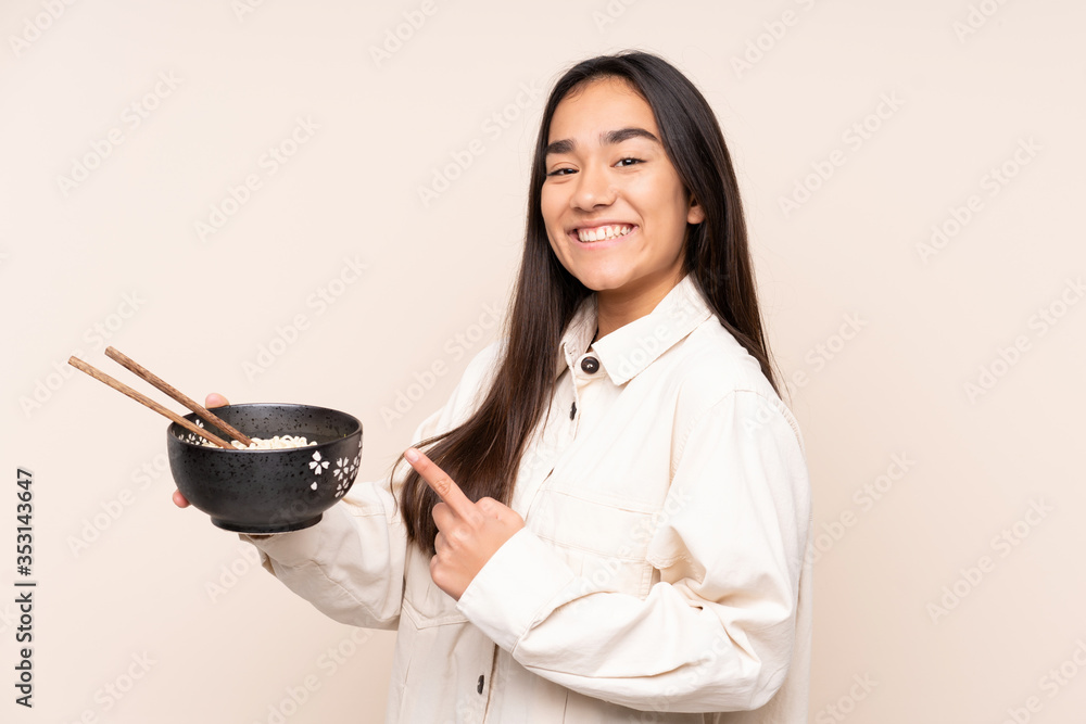 Young Indian woman isolated on beige background and pointing it while holding a bowl of noodles with chopsticks Young Indian woman isolated on beige background and pointing it while holding a bowl of noodles with chopsticks