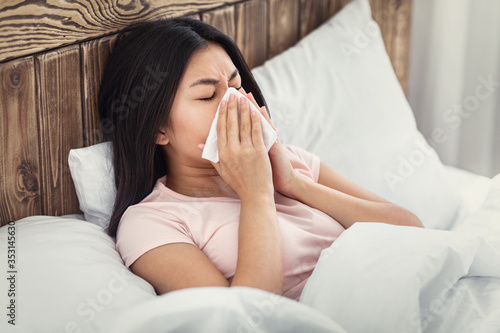 Sick Chinese Woman Blowing Nose In Tissue Lying In Bed