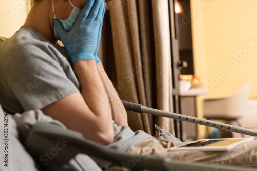 Selective focus of upset man in medical mask and latex gloves covering face near crutches and digital tablet on bed