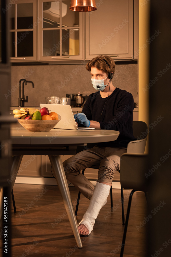 Selective focus of disabled man in medical mask using headset and laptop near fruits on kitchen table Selective focus of disabled man in medical mask using headset and laptop near fruits on kitchen table