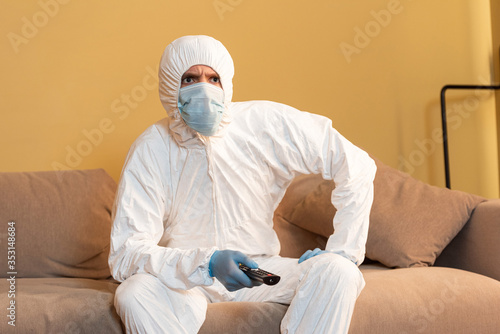 Thoughtful man in hazmat suit and medical mask watching tv on sofa