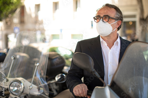 Uomo cinquantenne in giacca con mascherina protettiva modello kn95 ha appena parcheggiato il su scooter
 in un parcheggio in centro città  photo