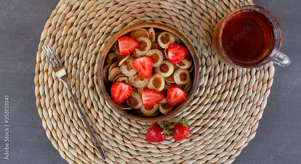 Tiny pancake cereal mini pancakes in a wooden bowl with strawberries, tea and fork on a concrete background. Top view. Copy space Tiny pancake cereal mini pancakes in a wooden bowl with strawberries, tea and fork on a concrete background. Top view. Copy space
