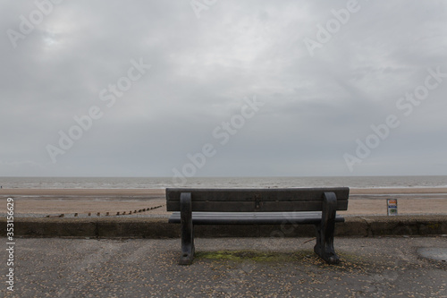 Image of a lonely black bench in England facing the sea with a long sand patch and stretch go pebbles going towards the water ready for someone depressed to sit on the lonely empty chair