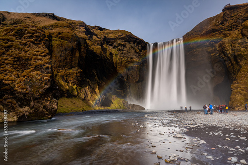 Skogafoss waterfall in South Iceland with a beautiful rainbow. Tourists admiring the waterfall. Popular and famous tourist attraction.