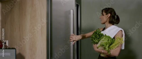 Sporty fit female dancing in the kitchen while picking ingredients for green juice cocktail from a fridge. Shot with 2x Anamorphic lens photo