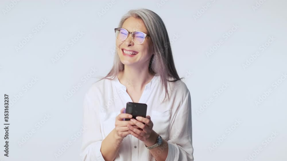A laughing happy old mature woman with long gray hair wearing glasses is using her smartphone standing isolated over white background in studio A laughing happy old mature woman with long gray hair wearing glasses is using her smartphone standing isolated over white background in studio