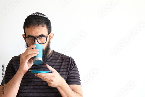 Bearded Jewish holding a cup of wine for the Kidush on Shabbat. The man is standing in front of a white background with a serious expression
