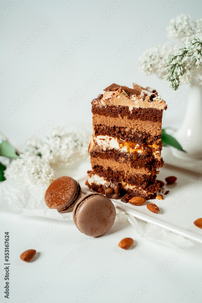 Chocolate cake decorated with various cookies and nuts on a glass plate and macaroons among white flowers of lilac and green leaves. Food photography. Advertising and commercial close up design. Chocolate cake decorated with various cookies and nuts on a glass plate and macaroons among white flowers of lilac and green leaves. Food photography. Advertising and commercial close up design.
