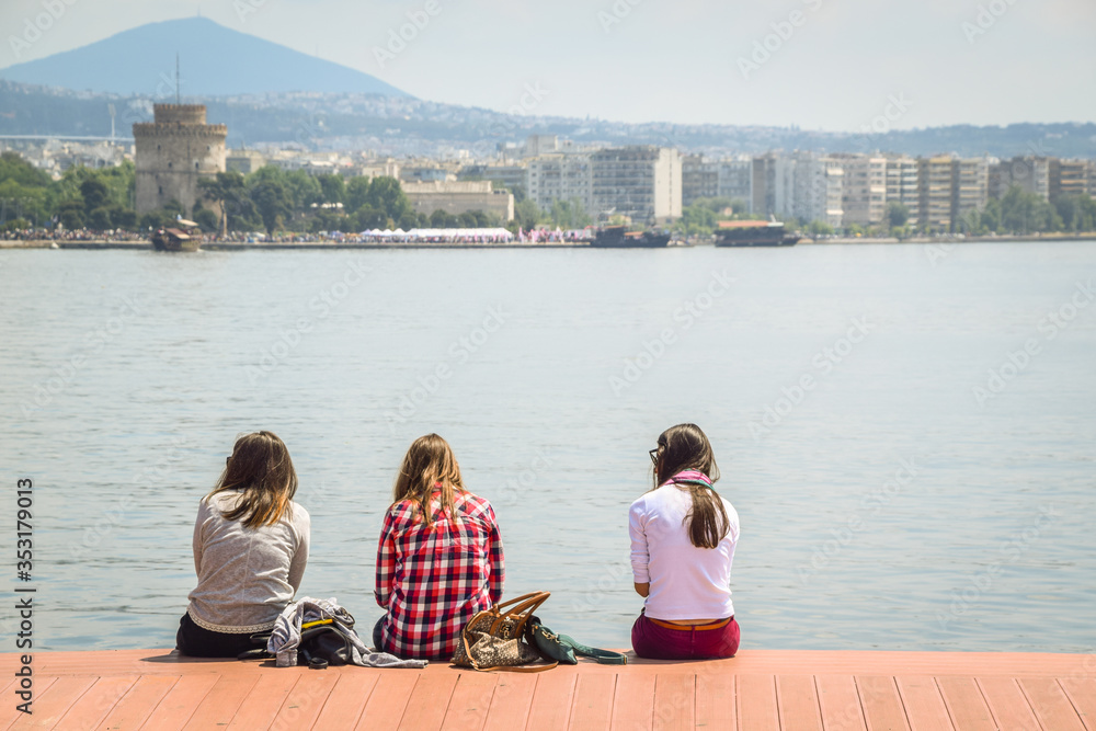 Three Teenagers Girls Friends Sitting On The Wooden Dock And Enjoying The City View In Front Of The Sea in Thessaloniki city in Greece, Europe.